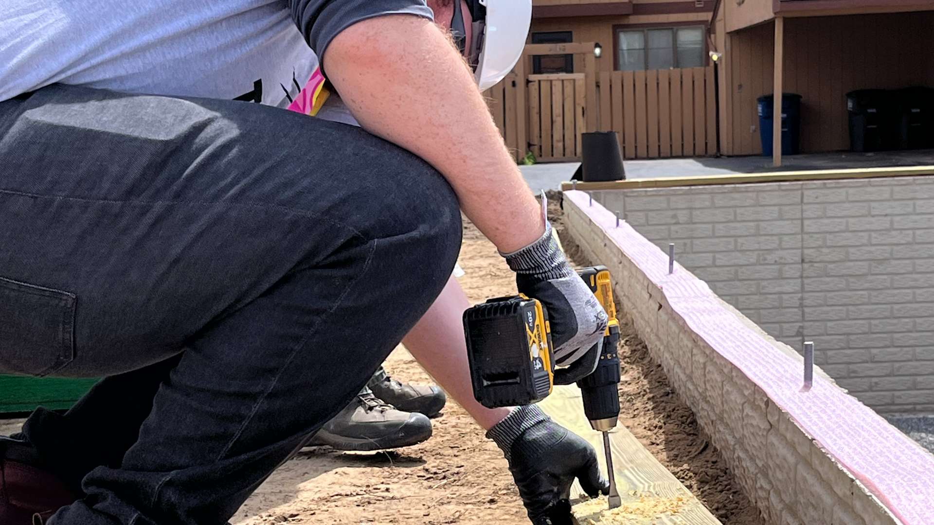 Volunteer wearing a white hard hat and black gloves using a power drill to secure a wooden plank on a foundation edge at an outdoor building site.