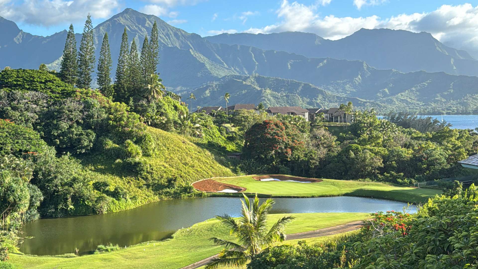 Scenic golf course with pond and palm trees surrounded by lush greenery and mountains under a bright blue sky.