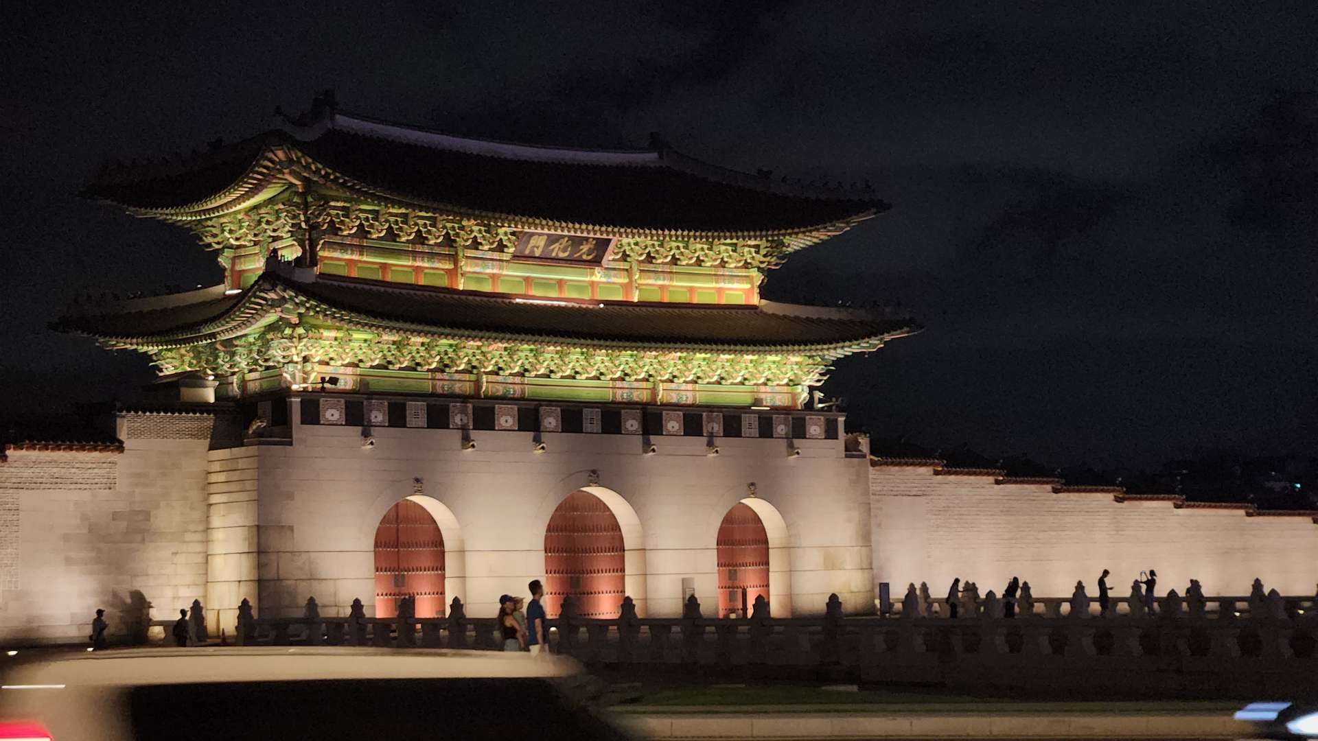 Illuminated traditional Korean palace gate at night with ornate green and red roof details and stone walls.