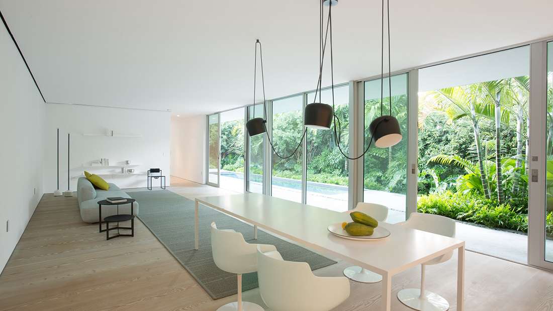 Minimalist open-concept living and dining area with floor-to-ceiling sliding glass doors overlooking lush tropical garden, featuring white dining table with modern chairs, black pendant lights, light wood flooring, and sleek gray sofa.