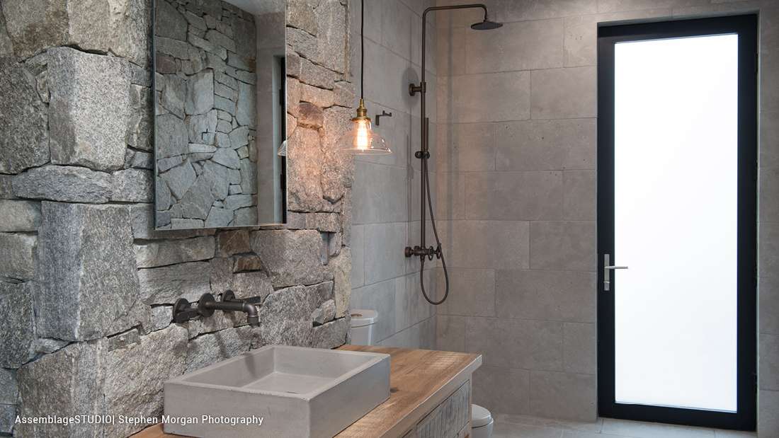 Luxury bathroom with stone accent wall, concrete vanity with rectangular sink, glass pendant light, and walk-in shower featuring matte black fixtures and frosted glass door.