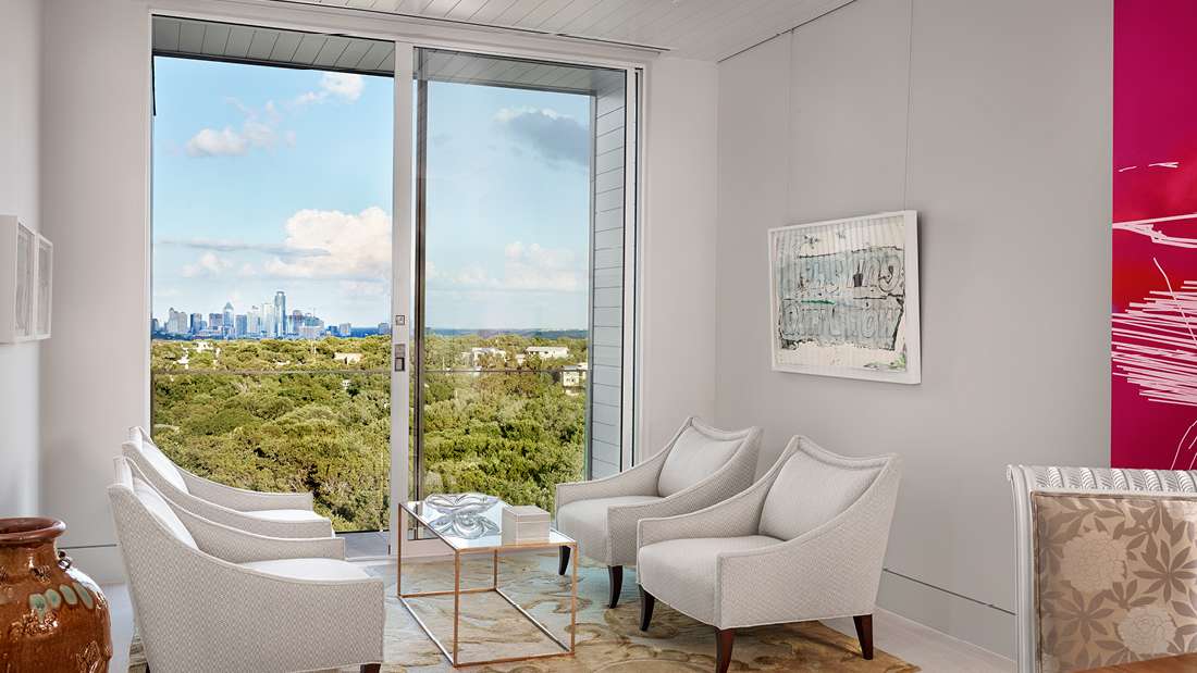 Bright modern sitting area with floor-to-ceiling glass doors offering city skyline views, white upholstered armchairs, and contemporary wall art.