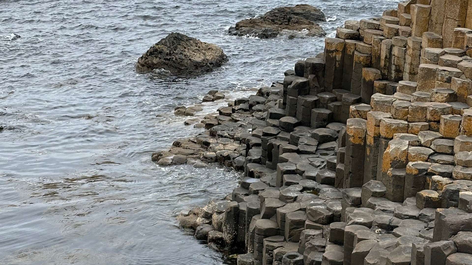 Hexagonal basalt columns along rocky shoreline with ocean waves and scattered boulders in the water.