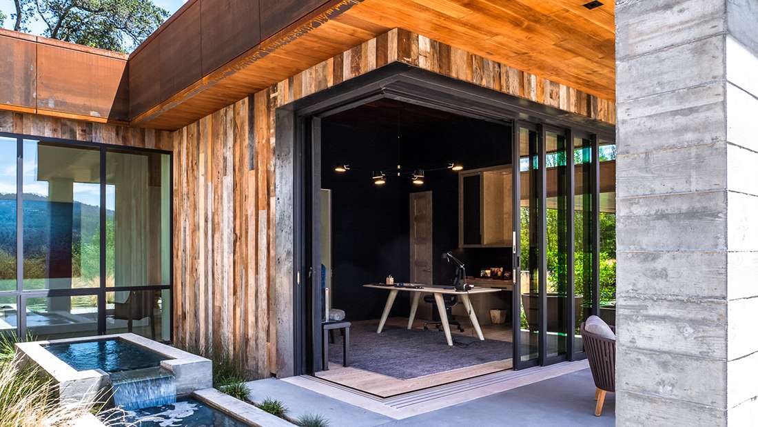 Modern home office with folding glass doors opening to outdoor patio, rustic wood siding, concrete columns, natural wood ceiling, and rectangular water feature surrounded by greenery.