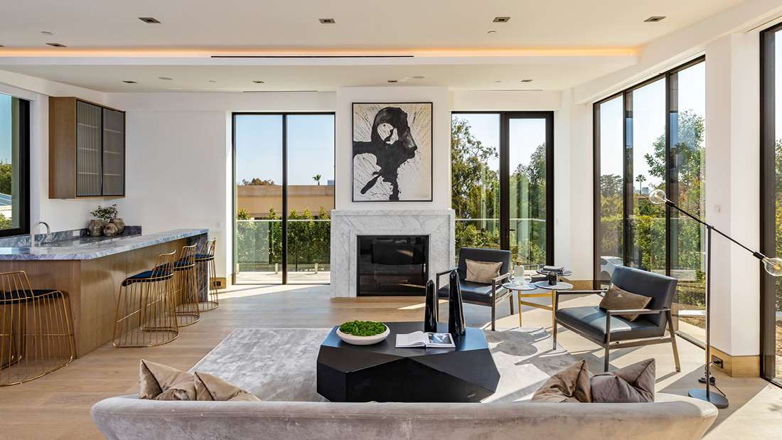 Luxury open-concept living room with floor-to-ceiling glass doors, marble fireplace, abstract black-and-white artwork, modern gray sofa, and kitchen bar with marble countertop and gold barstools.