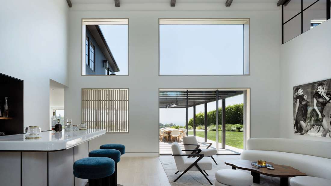 Bright modern living space with high ceilings, large windows, stylish bar area with blue stools, and sliding glass doors opening to a covered patio and green lawn.