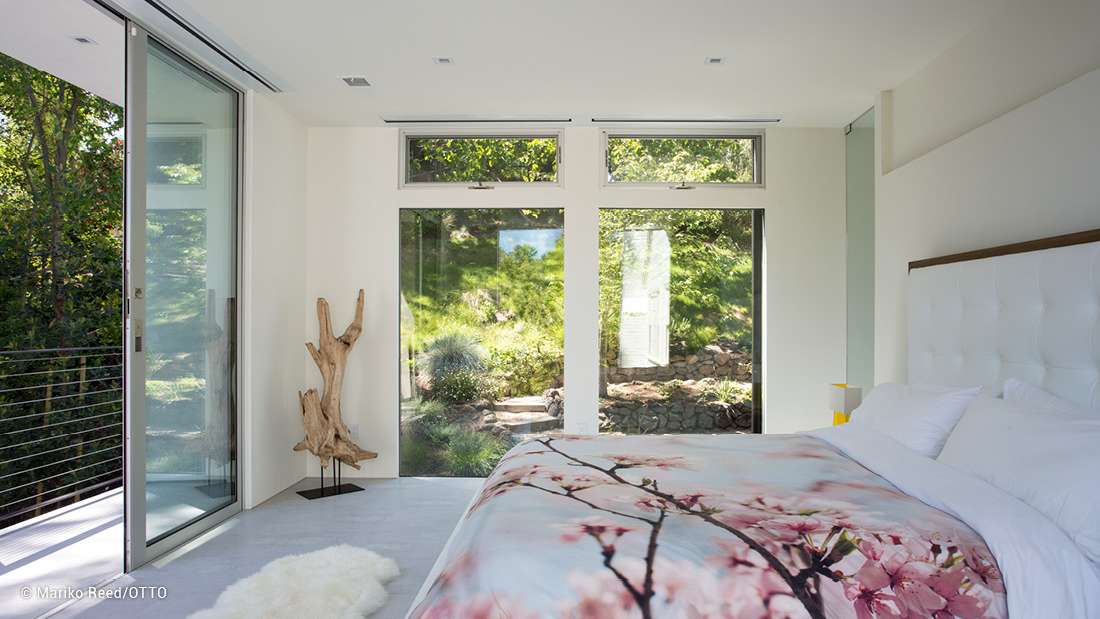 Bright modern bedroom with large sliding glass doors, minimalist white walls, floral-patterned bedding, and views of a lush green garden.