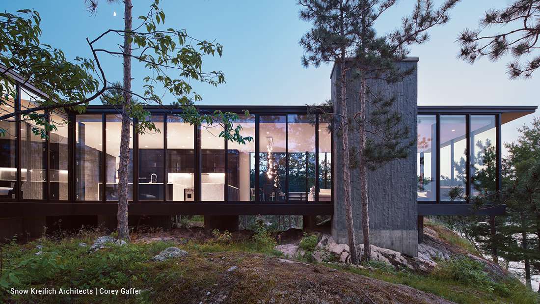 Contemporary lakefront home with elevated design, floor-to-ceiling glass walls, black-framed windows, and cantilevered structure surrounded by pine trees and rocky terrain.