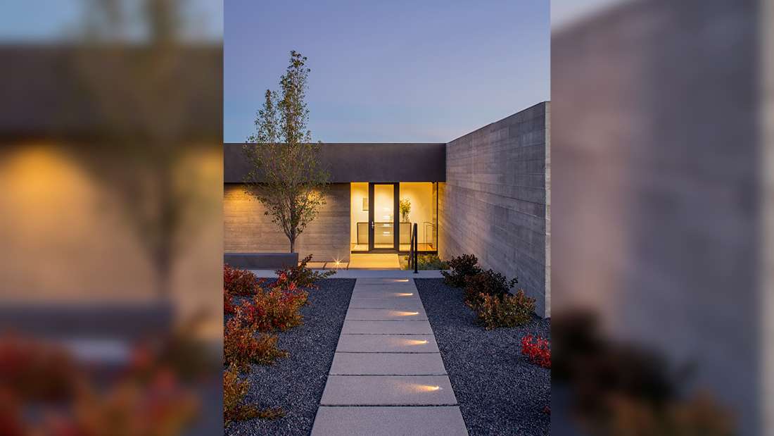 Minimalist modern home entrance with concrete walls, recessed lighting, and straight pathway lined with square pavers leading to glass front door, surrounded by desert landscaping and colorful shrubs at dusk.