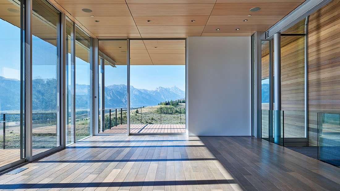 Minimalist modern interior with floor-to-ceiling glass walls, sliding glass doors, light wood ceiling panels, and wide-plank hardwood flooring overlooking panoramic mountain views.