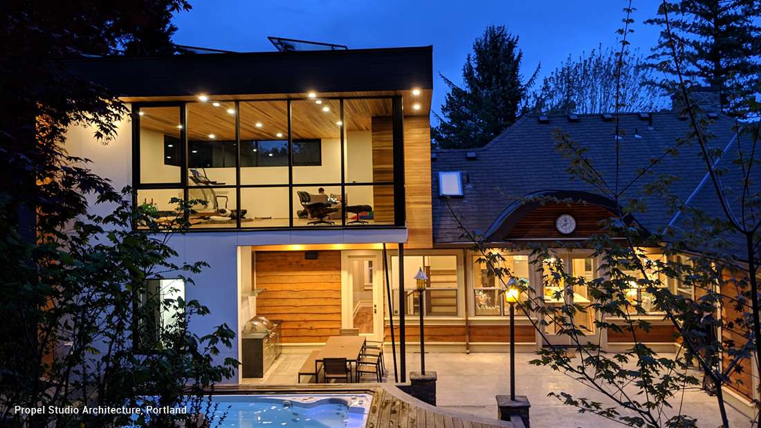 Contemporary home exterior at dusk featuring two-story glass walls, wood-paneled ceiling with recessed lighting, outdoor dining area with built-in grill, and elevated hot tub surrounded by trees.