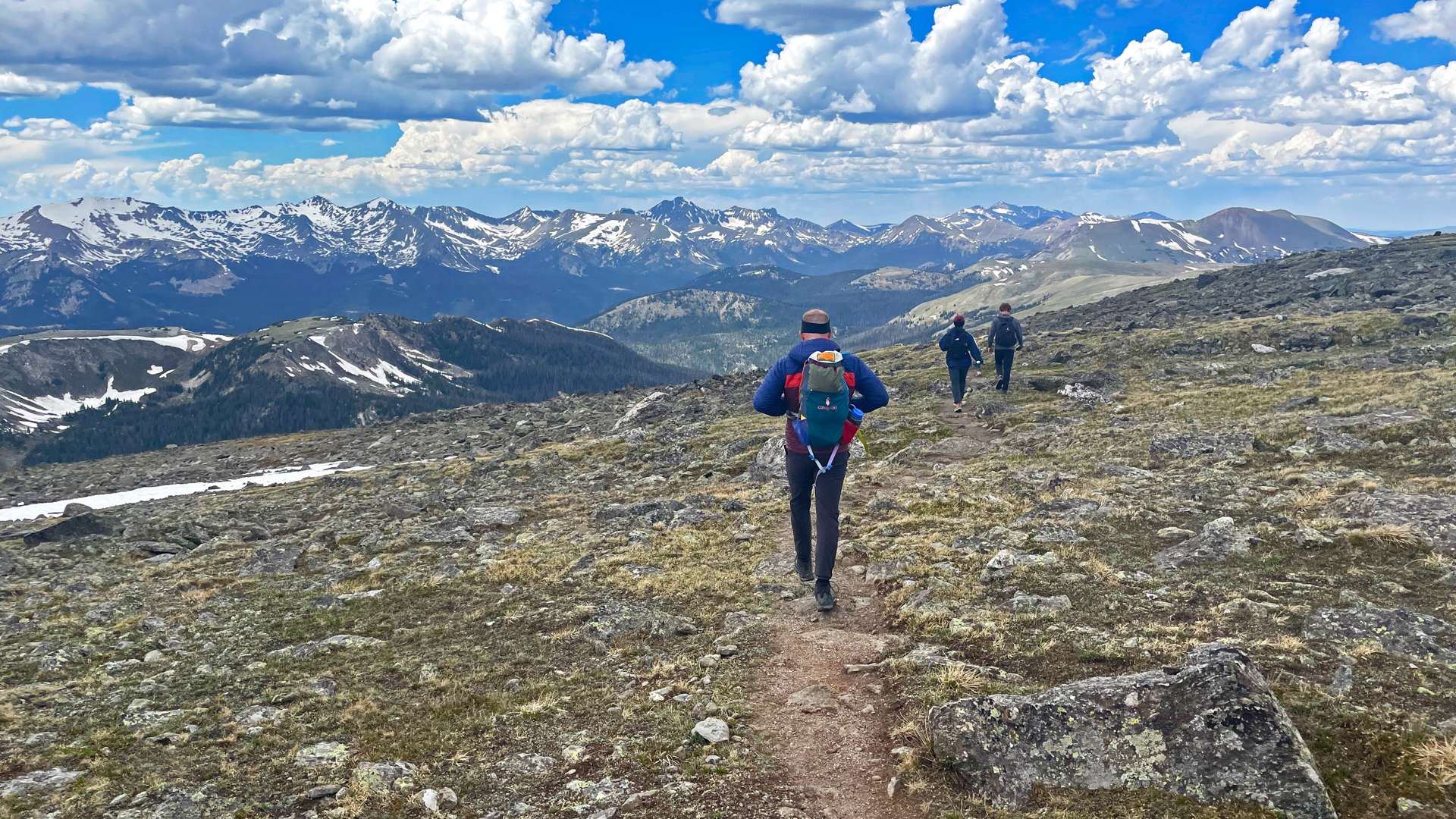 Three hikers walking along a narrow dirt trail on a rocky alpine ridge, with snow-capped mountain peaks and dramatic clouds in the distance under a bright blue sky.