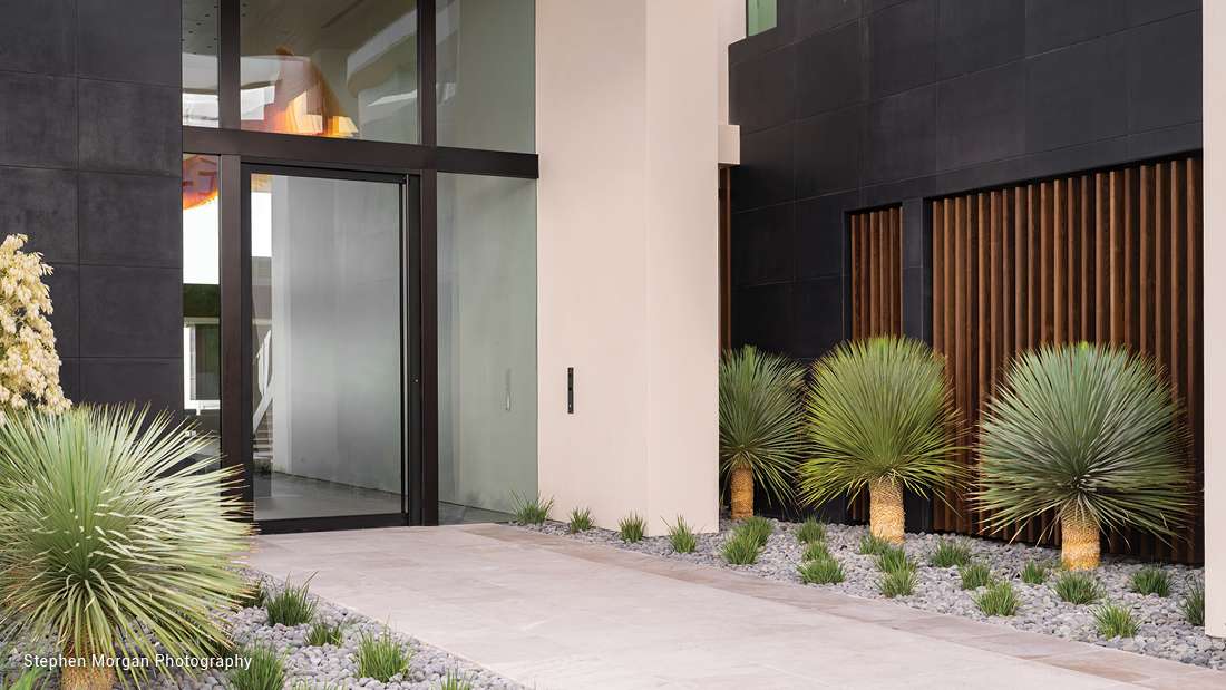 Modern home entryway with black-framed glass pivot door, floor-to-ceiling windows, beige stucco walls, and minimalist desert landscaping featuring agave plants and gray gravel.