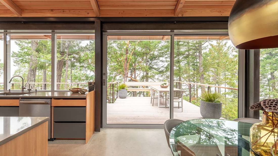 Modern kitchen and dining area with wood. cabinetry, glass table, and sliding glass doors opening to a forest-view deck with outdoor seating.