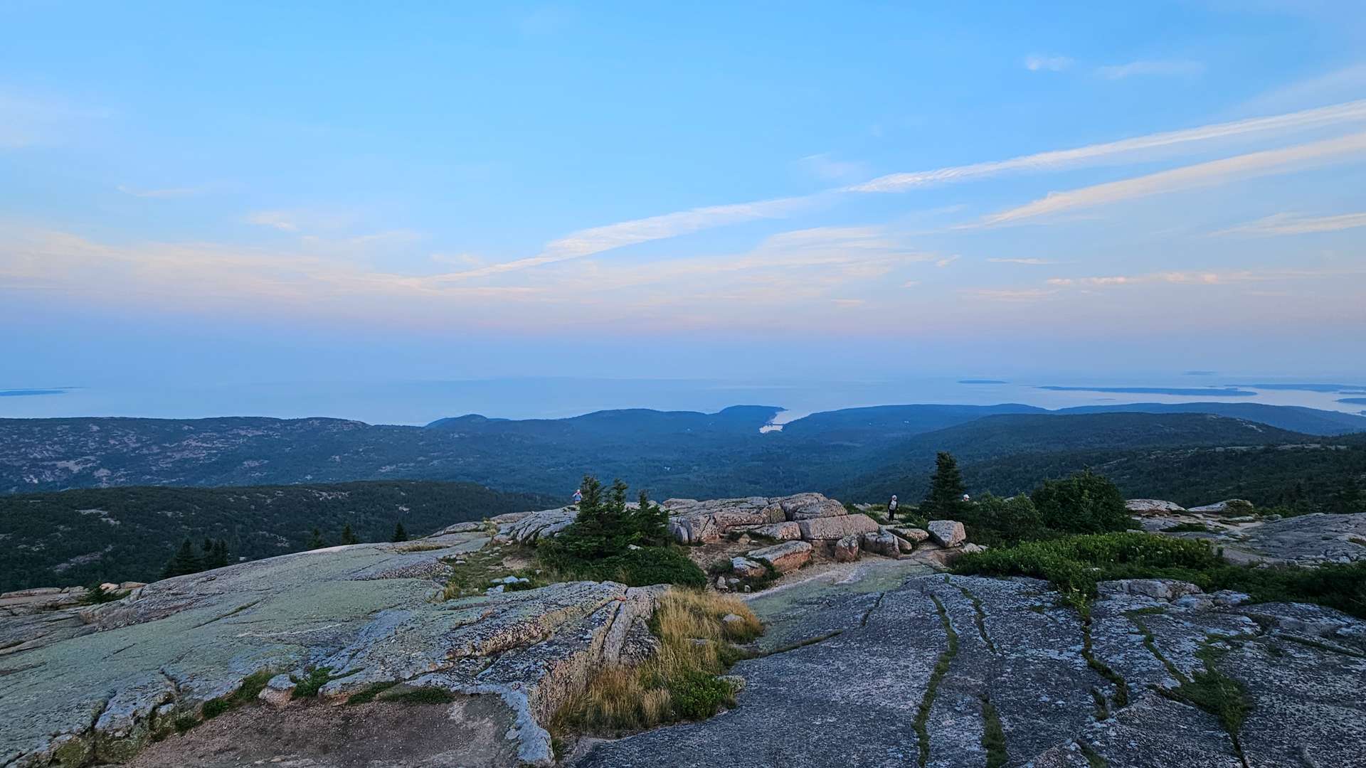 Rocky mountain summit overlooking forested hills and distant ocean under a clear blue sky.