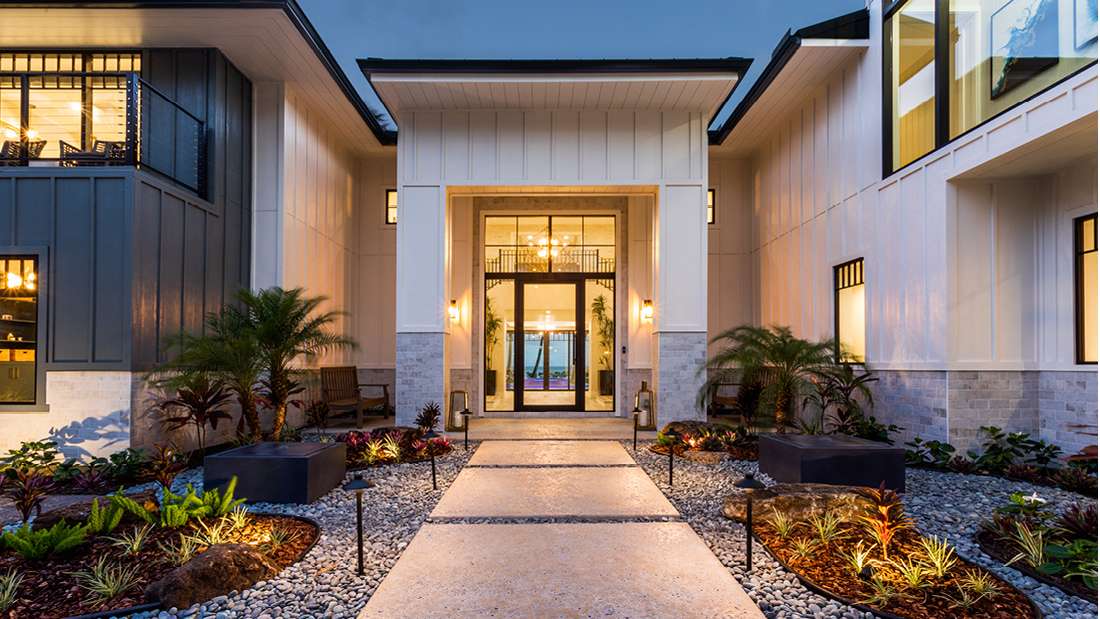 Luxury modern home exterior with symmetrical design, white vertical siding, black-framed glass entry door, and landscaped walkway featuring gravel beds, tropical plants, and accent lighting.