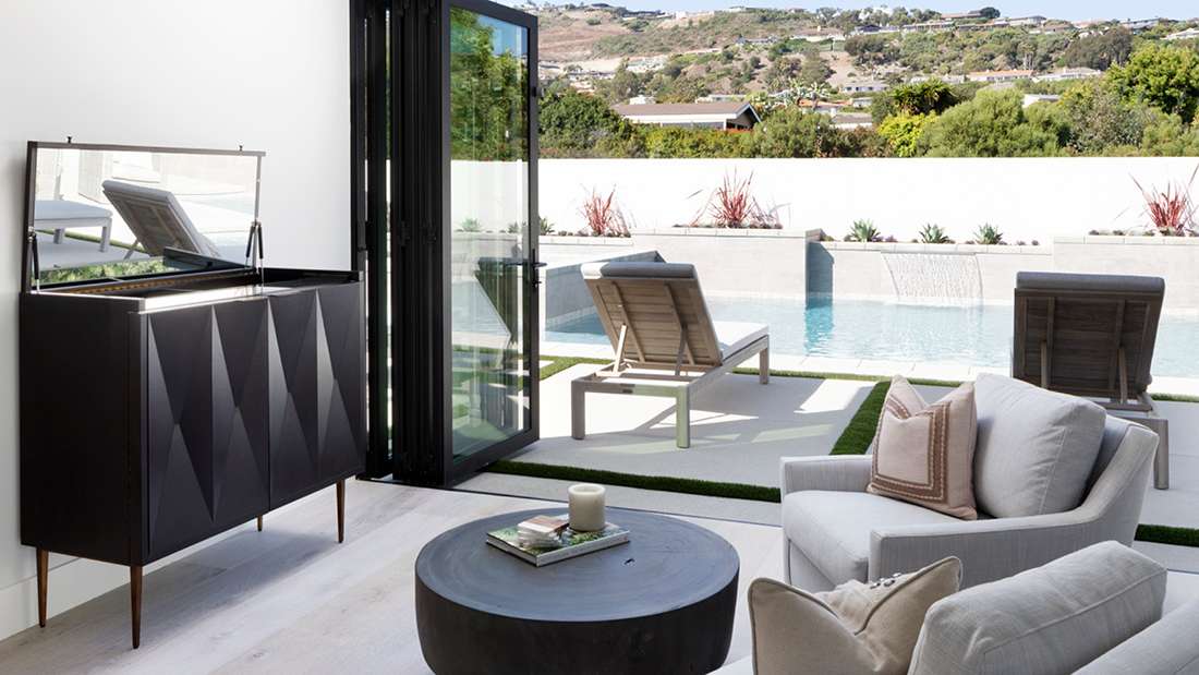 Modern living area with light gray armchair and round black coffee table, folding glass doors opening to outdoor pool deck with two lounge chairs and waterfall feature, surrounded by lush greenery and hillside views.