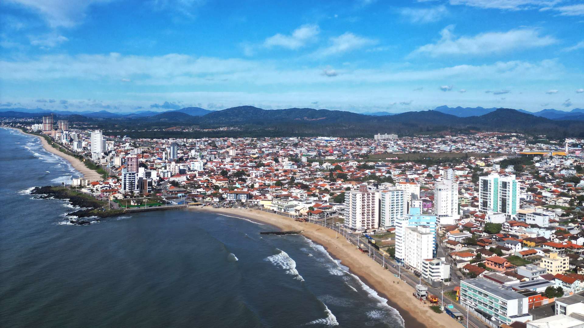   “Aerial view of a coastal city with sandy beaches, high-rise buildings, and distant mountains under a bright blue sky.