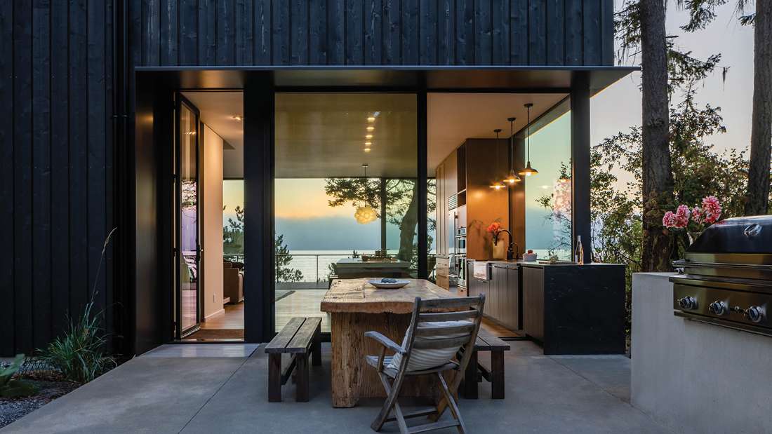 Modern outdoor dining area with rustic wooden table and benches, surrounded by black vertical siding, floor-to-ceiling glass walls, and open kitchen featuring pendant lights and stainless steel appliances overlooking ocean sunset.