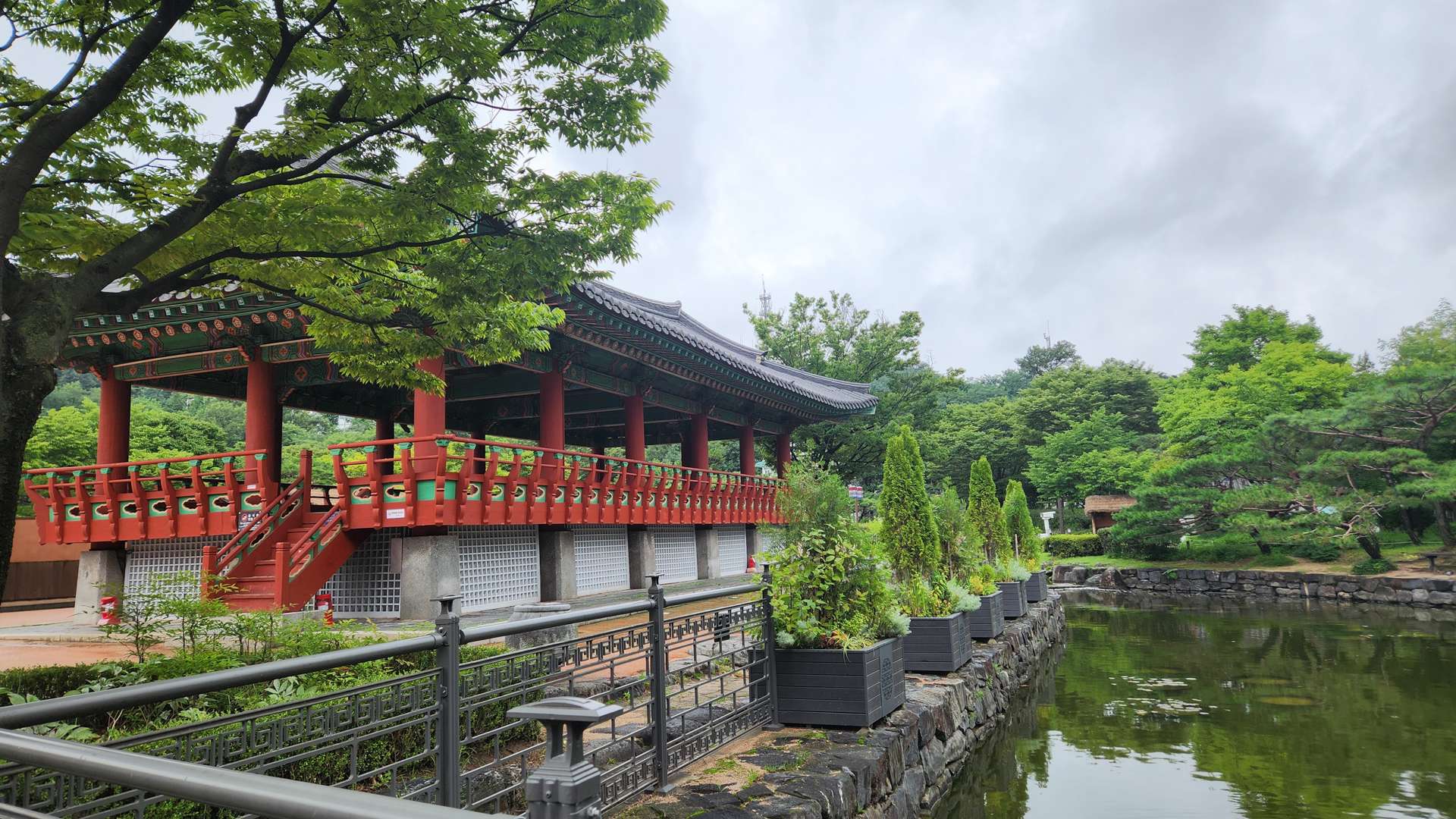 Traditional Korean pavilion with red columns beside a calm pond surrounded by lush green trees.