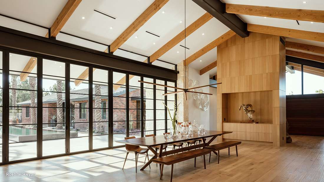 Luxury dining room with vaulted ceiling featuring exposed wooden beams, floor-to-ceiling glass doors, long wooden dining table with matching benches, and built-in light wood cabinetry with modern decor.