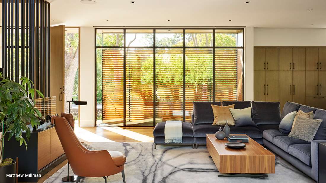 Modern living room with floor-to-ceiling glass doors featuring horizontal wooden slats, navy sectional sofa with assorted throw pillows, tan leather accent chair, wooden coffee table, and built-in cabinetry.