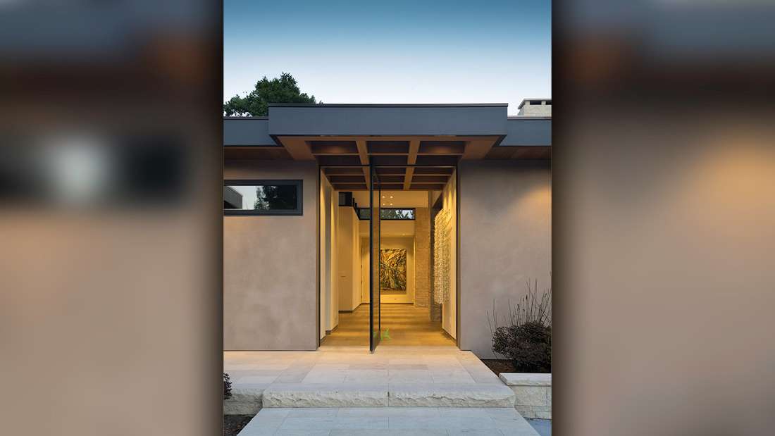 Modern home entryway with pivot glass door, flat roof design featuring geometric overhang, beige stucco walls, and minimalist landscaping with stone pavers and recessed lighting.