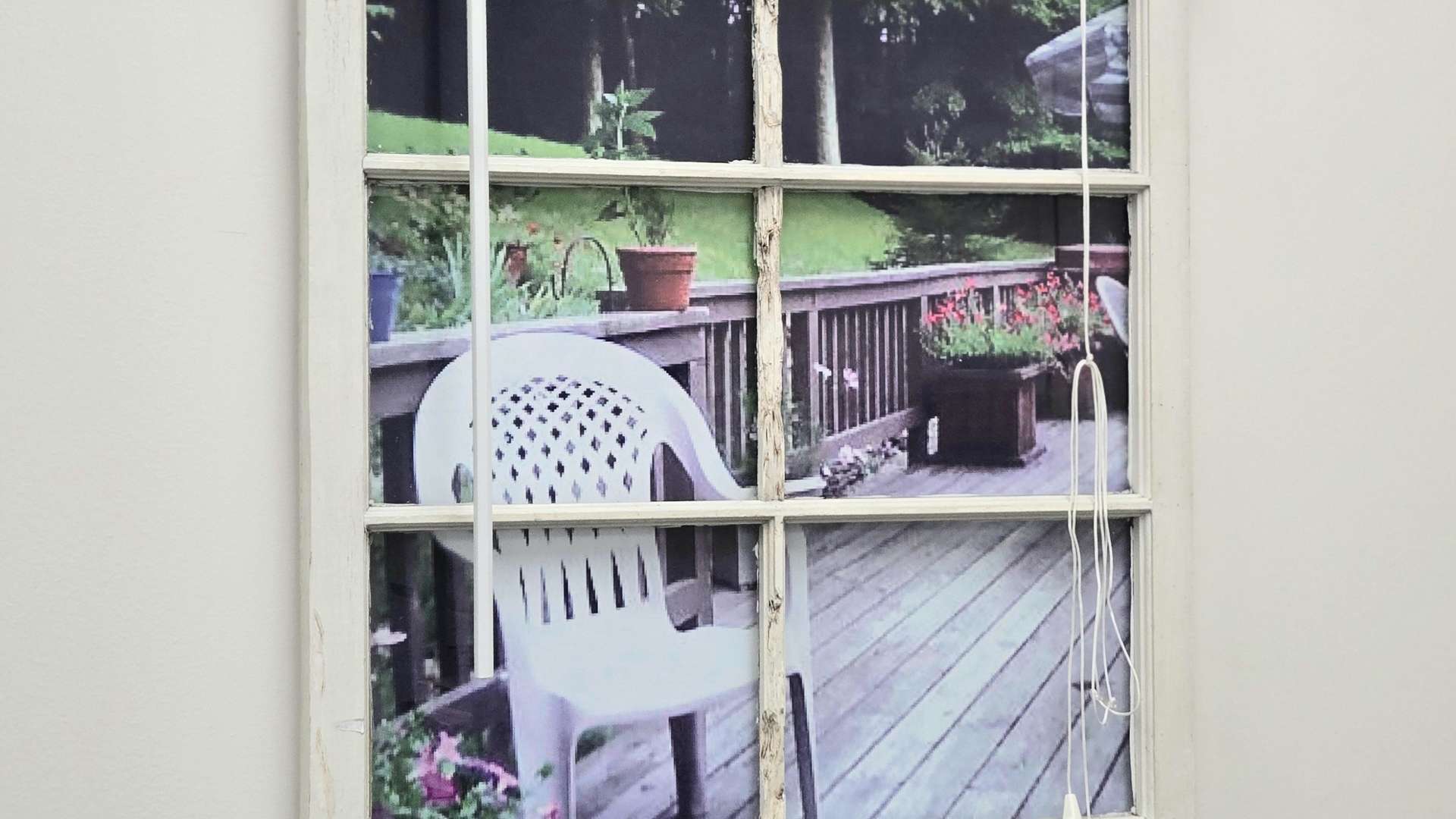  Decorative window frame with blinds showing a printed photo of a patio with chair, plants, and wooden railing.