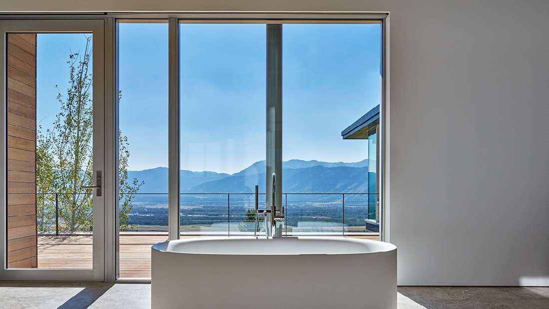 Luxury bathroom with freestanding oval bathtub positioned in front of floor-to-ceiling glass doors and windows, offering panoramic mountain and valley views from an outdoor deck.