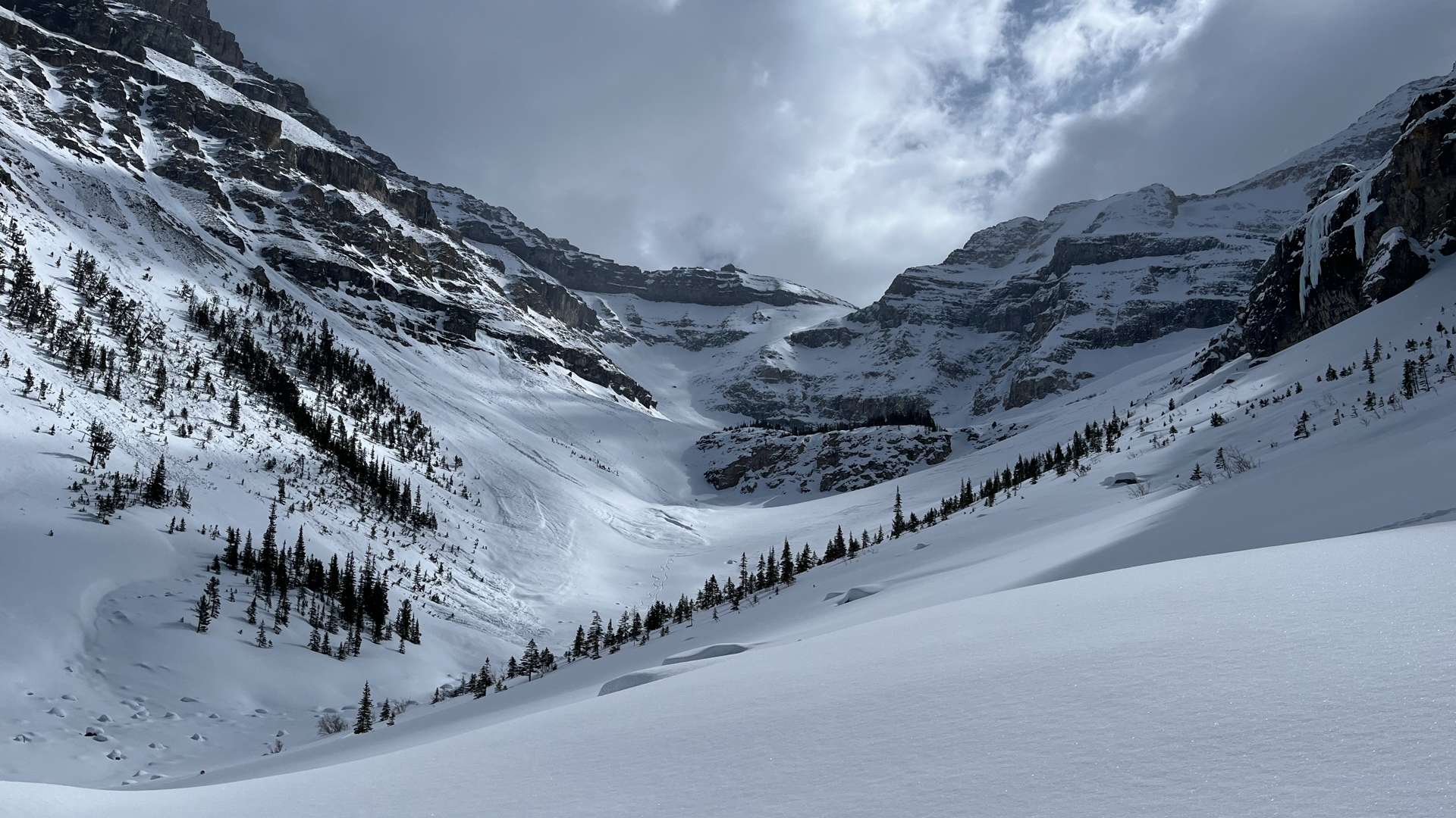 Snow-covered alpine valley surrounded by steep rocky mountains under a dramatic cloudy sky.