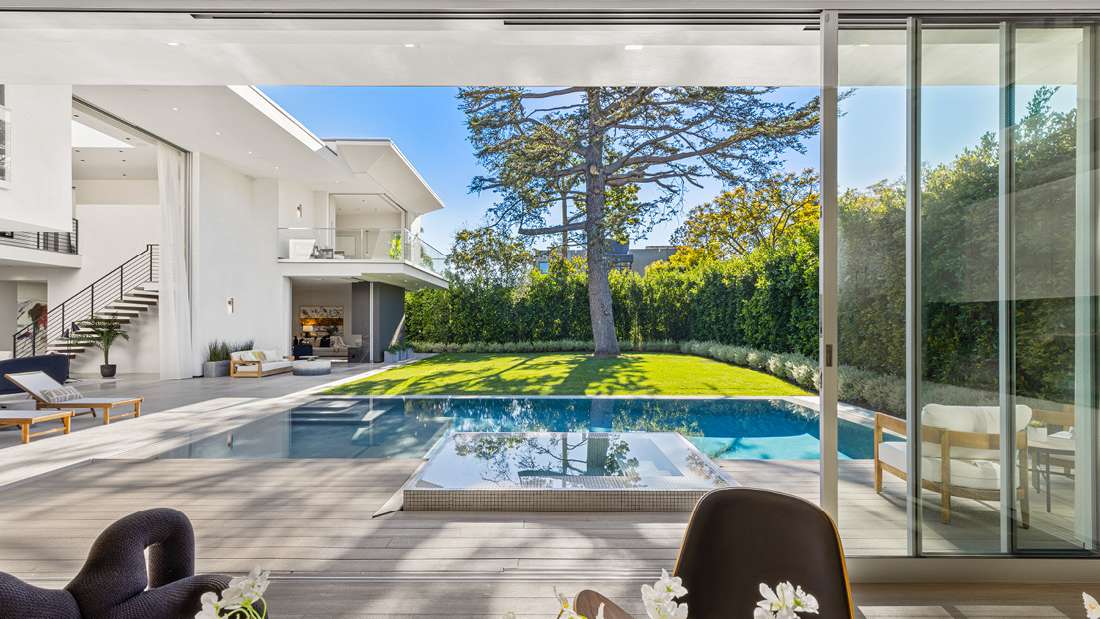 Luxury modern home with open-concept design featuring sliding glass doors opening to a backyard with swimming pool, integrated spa, wooden deck, and manicured lawn surrounded by tall hedges.