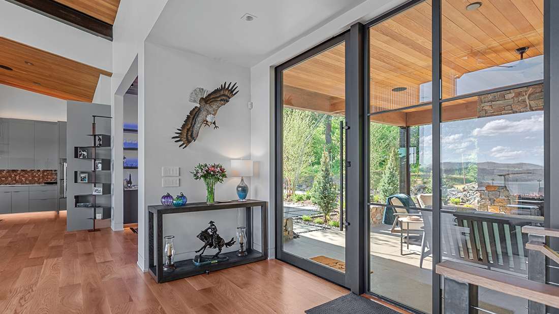 Modern home interior with glass double doors opening to a covered patio, natural wood ceiling panels, light hardwood flooring, and decorative console table featuring floral arrangement and wall-mounted eagle sculpture.