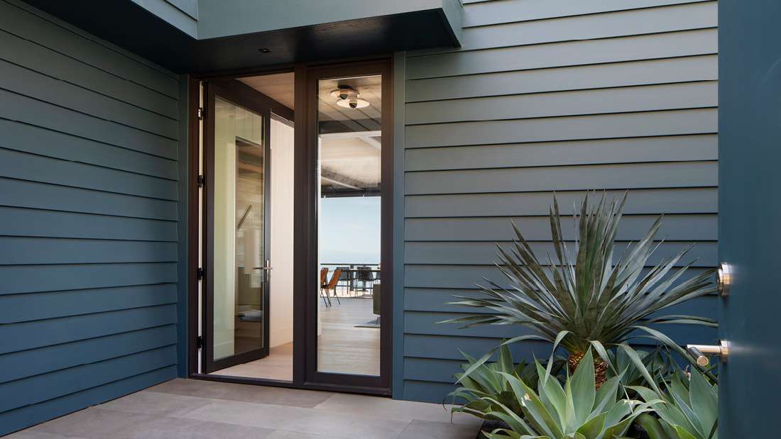 Modern home entryway with horizontal blue siding, black-framed glass door, and outdoor landscaping featuring agave plants on a tiled patio.