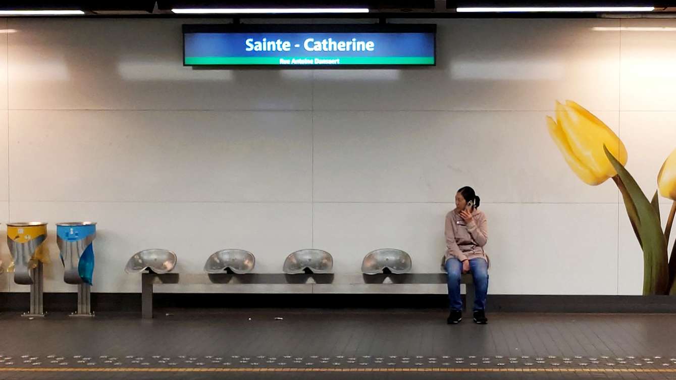 Person sitting on a bench at Sainte-Catherine metro station, talking on a phone, with large yellow tulip murals on the wall and recycling bins nearby.