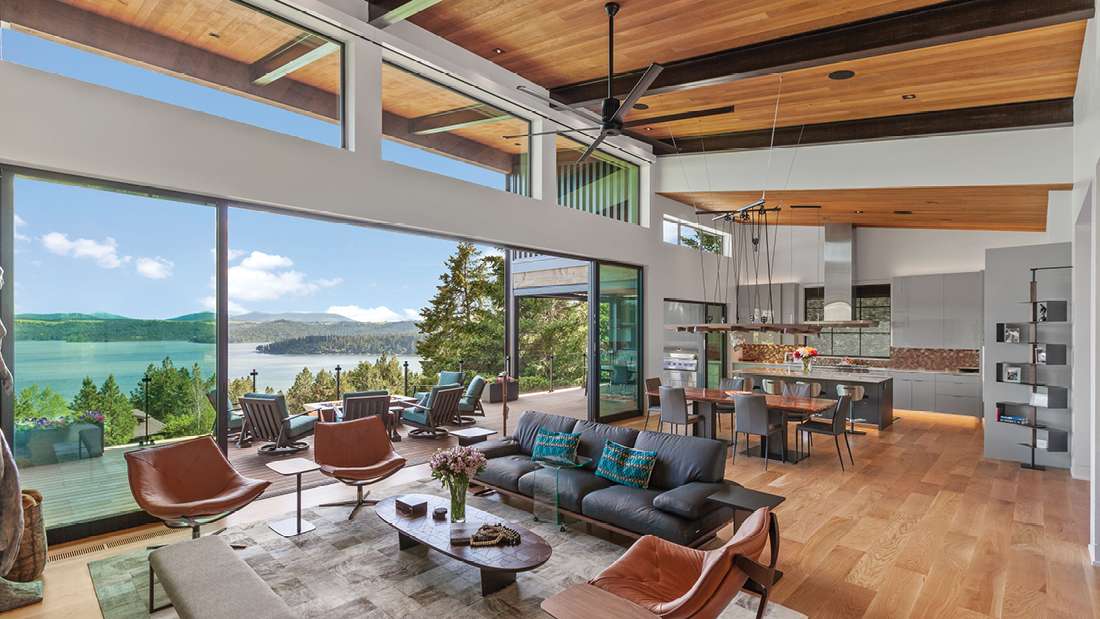 Luxury open-concept living and dining area with exposed wood ceiling beams, floor-to-ceiling glass walls, modern leather seating, and outdoor deck overlooking a scenic lake and forested hills.