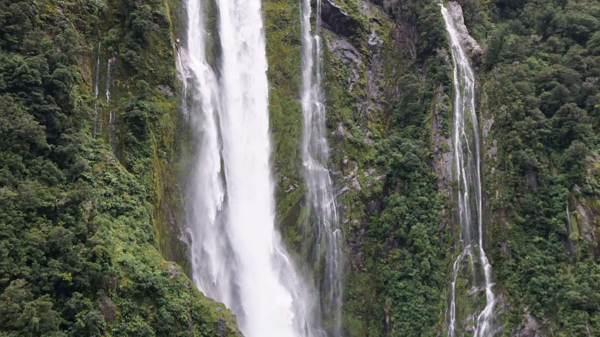   Tall waterfall cascading down lush green cliffs into a misty pool surrounded by dense forest vegetation.