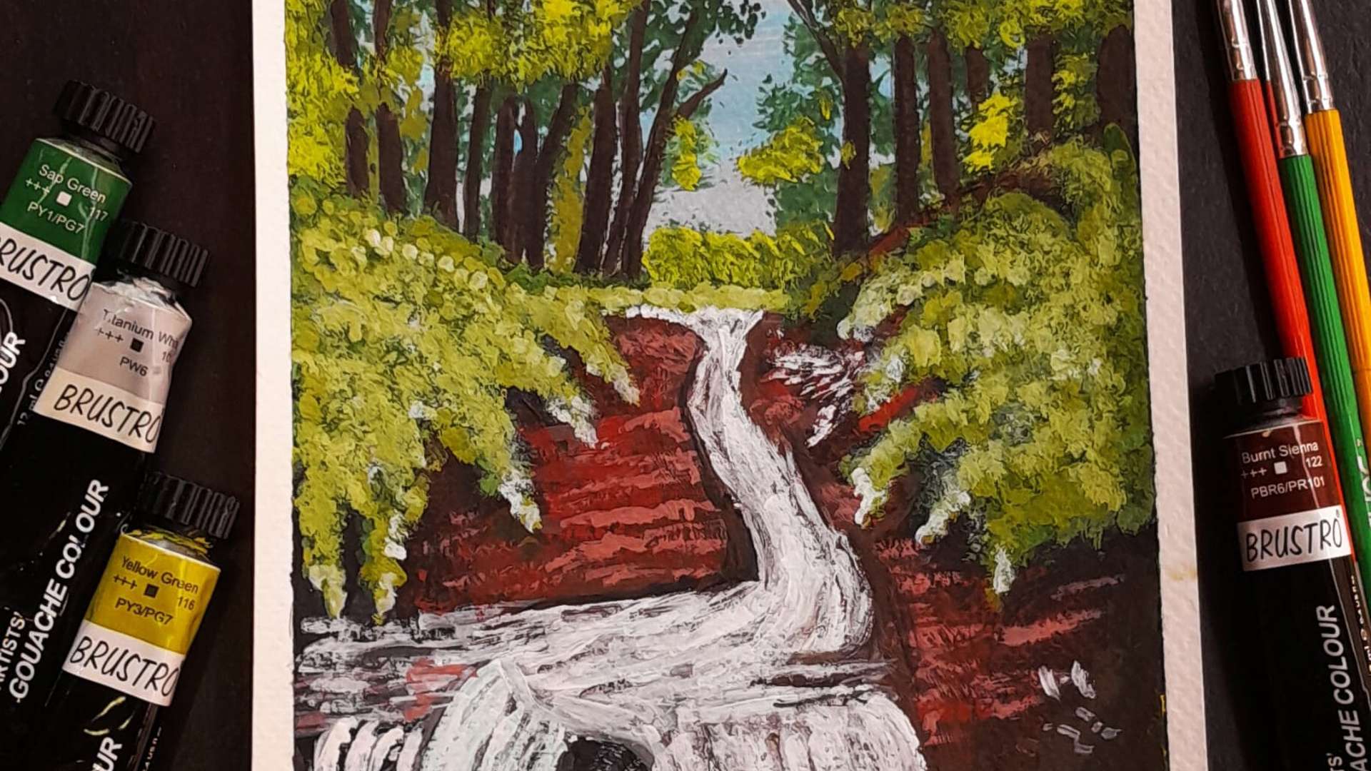   Acrylic painting of a forest waterfall with green trees, red soil, and paint tubes and brushes beside the artwork.