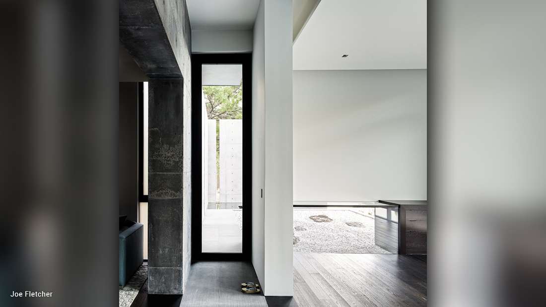 Minimalist modern hallway with narrow black-framed glass door, polished dark wood flooring, exposed concrete column, and adjacent open space featuring light gray walls and gravel garden visible through large window.