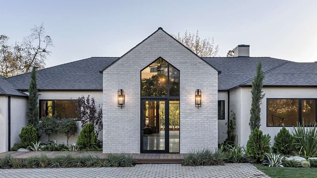 Modern farmhouse-style home exterior with white brick facade, steep gabled roof, black-framed glass entry door, symmetrical wall sconces, and landscaped front yard featuring shrubs and ornamental grasses.