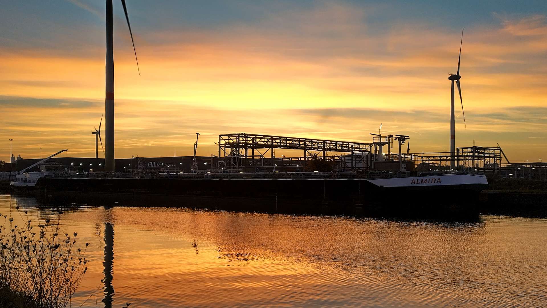 Wind turbines and industrial structures reflected in calm water at sunset with vibrant orange and blue sky.
