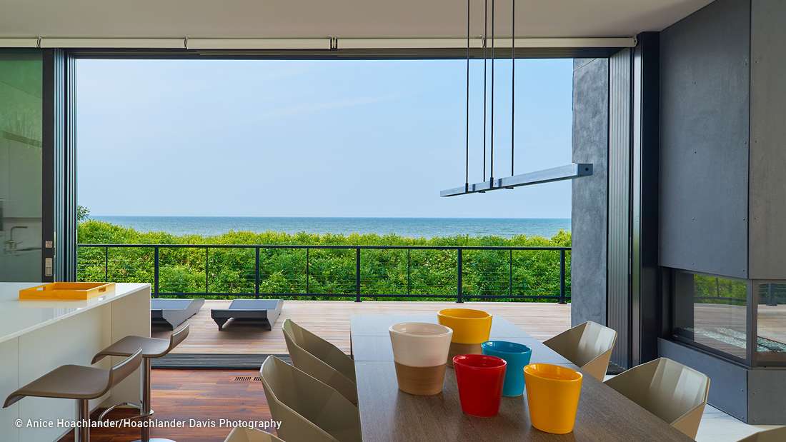 Modern dining area with large sliding glass doors opening to a deck overlooking ocean views, minimalist gray dining chairs, wooden table with colorful ceramic bowls, and sleek pendant light fixture.