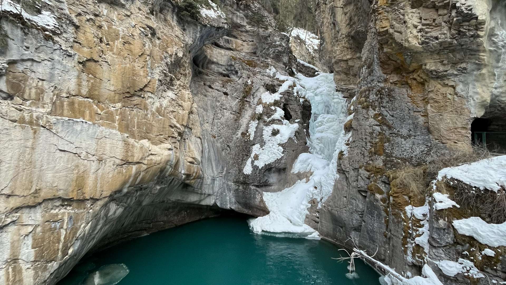 Frozen waterfall cascading into a turquoise pool surrounded by rugged canyon walls and patches of snow.