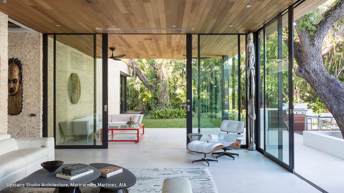 Modern indoor-outdoor living space with floor-to-ceiling sliding glass doors, natural wood ceiling panels, textured stone walls, and minimalist furniture including white lounge chairs and coffee table, opening to a lush tropical garden.