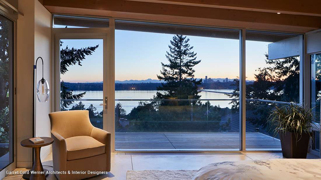 Modern bedroom with floor-to-ceiling glass doors opening to a balcony overlooking a lake and mountains at sunset, beige armchair with side table and pendant light, and minimalist decor with exposed wood ceiling beams.