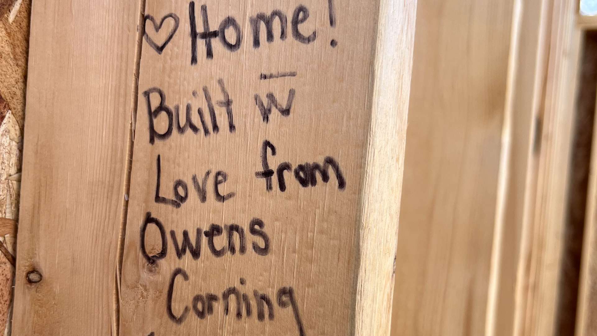   Close-up of a wooden wall stud with handwritten message that reads “Welcome Home! Built with love from Owens Corning Teams” and a smiley face drawn underneath.