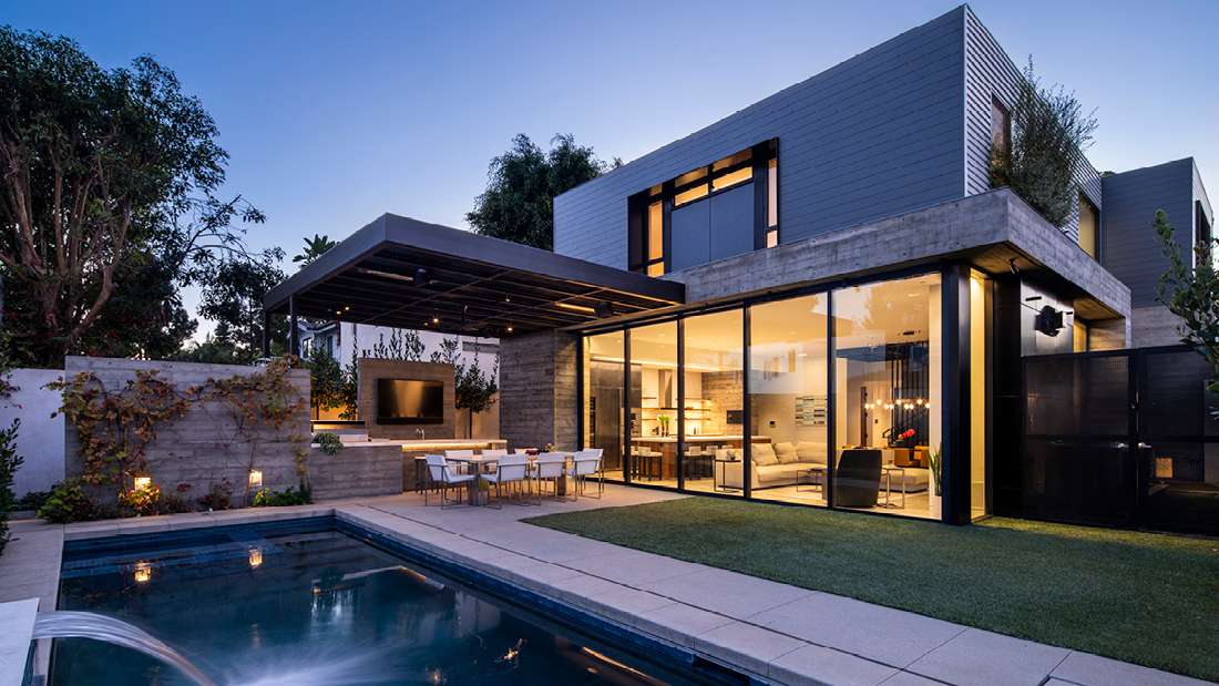 Luxury modern home exterior with floor-to-ceiling glass walls, open-concept living area, covered outdoor dining space with built-in TV, and rectangular swimming pool featuring water fountain jets.