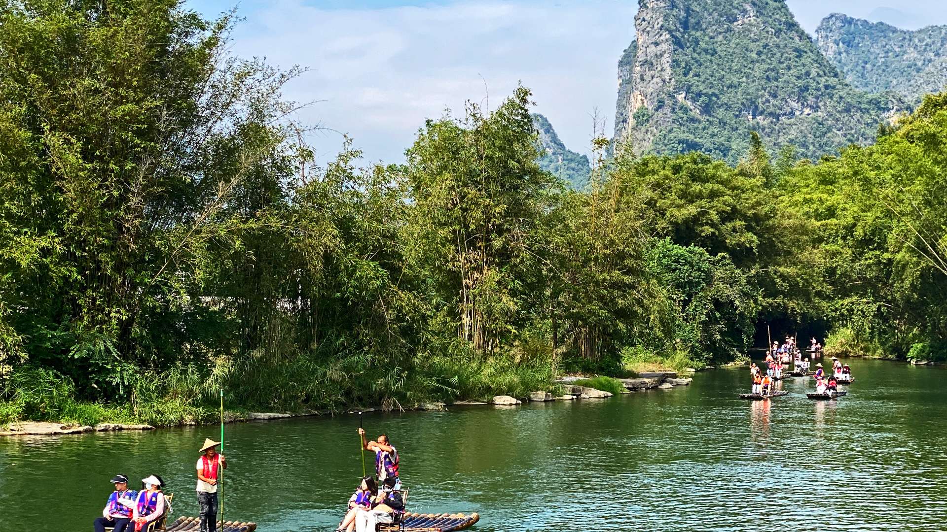 Tourists rafting on a calm green river surrounded by dense bamboo and karst mountains under a clear sky.