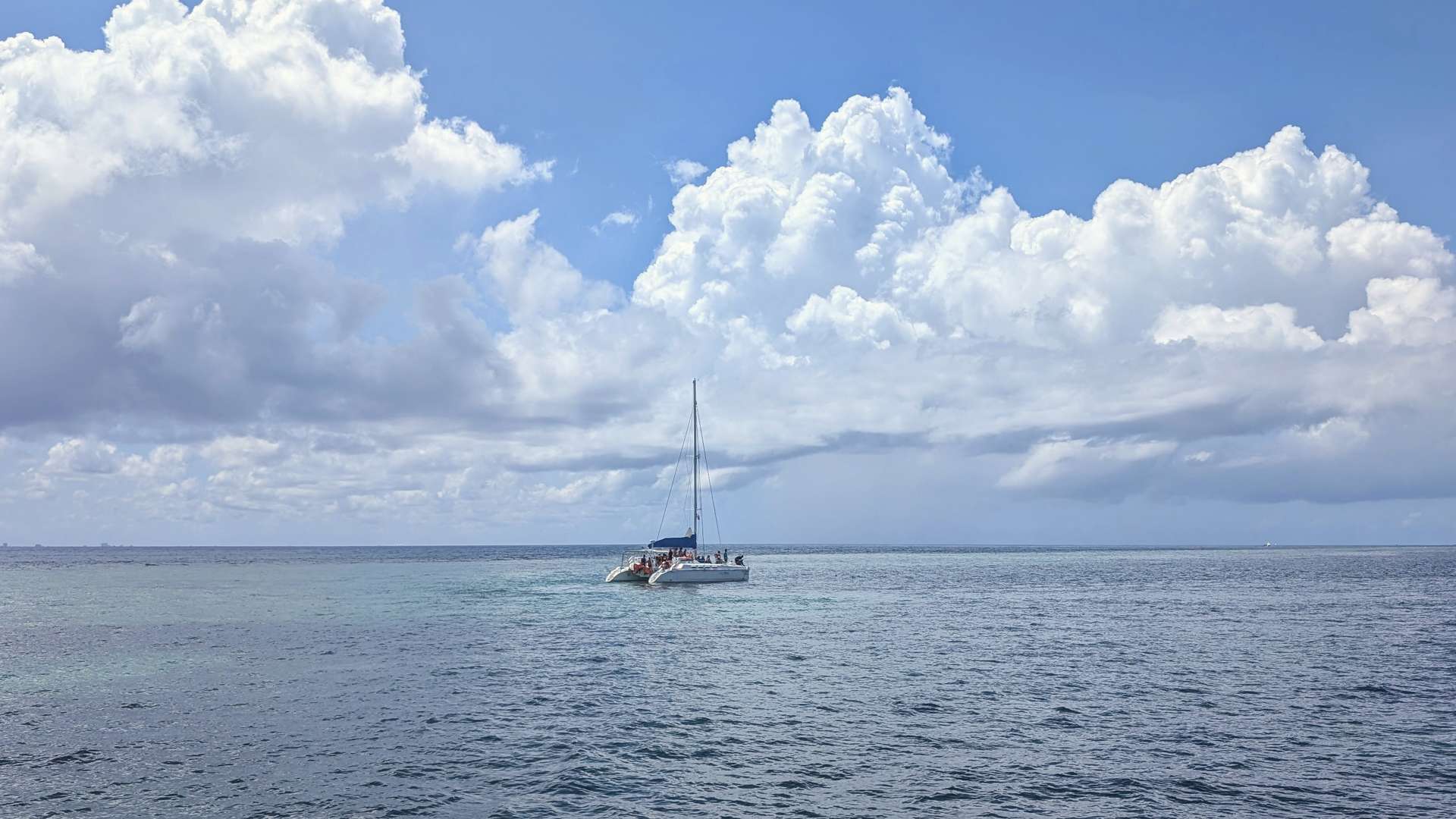White sailboat floating on calm blue ocean under a bright sky with large fluffy clouds.