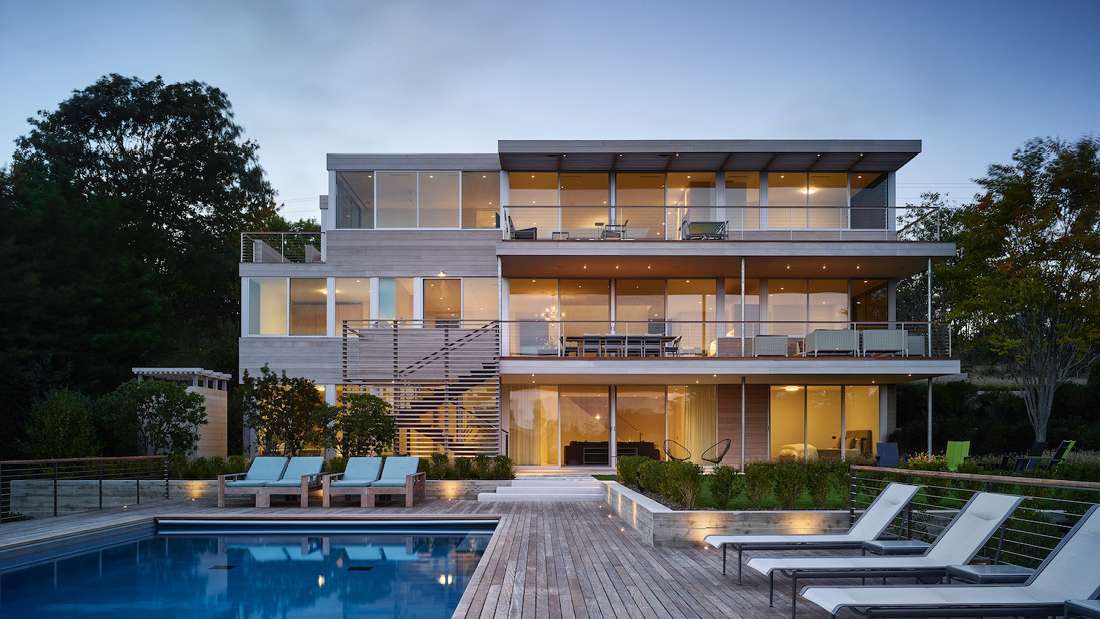 Modern three-story luxury home with expansive glass windows, multiple balconies, outdoor lounge chairs, and a large swimming pool surrounded by wooden deck.