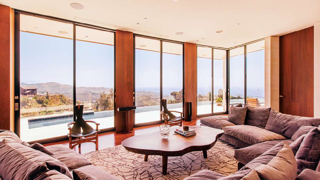 Luxury living room with floor-to-ceiling glass walls, panoramic mountain views, infinity pool outside, and plush sectional seating with wooden accents.